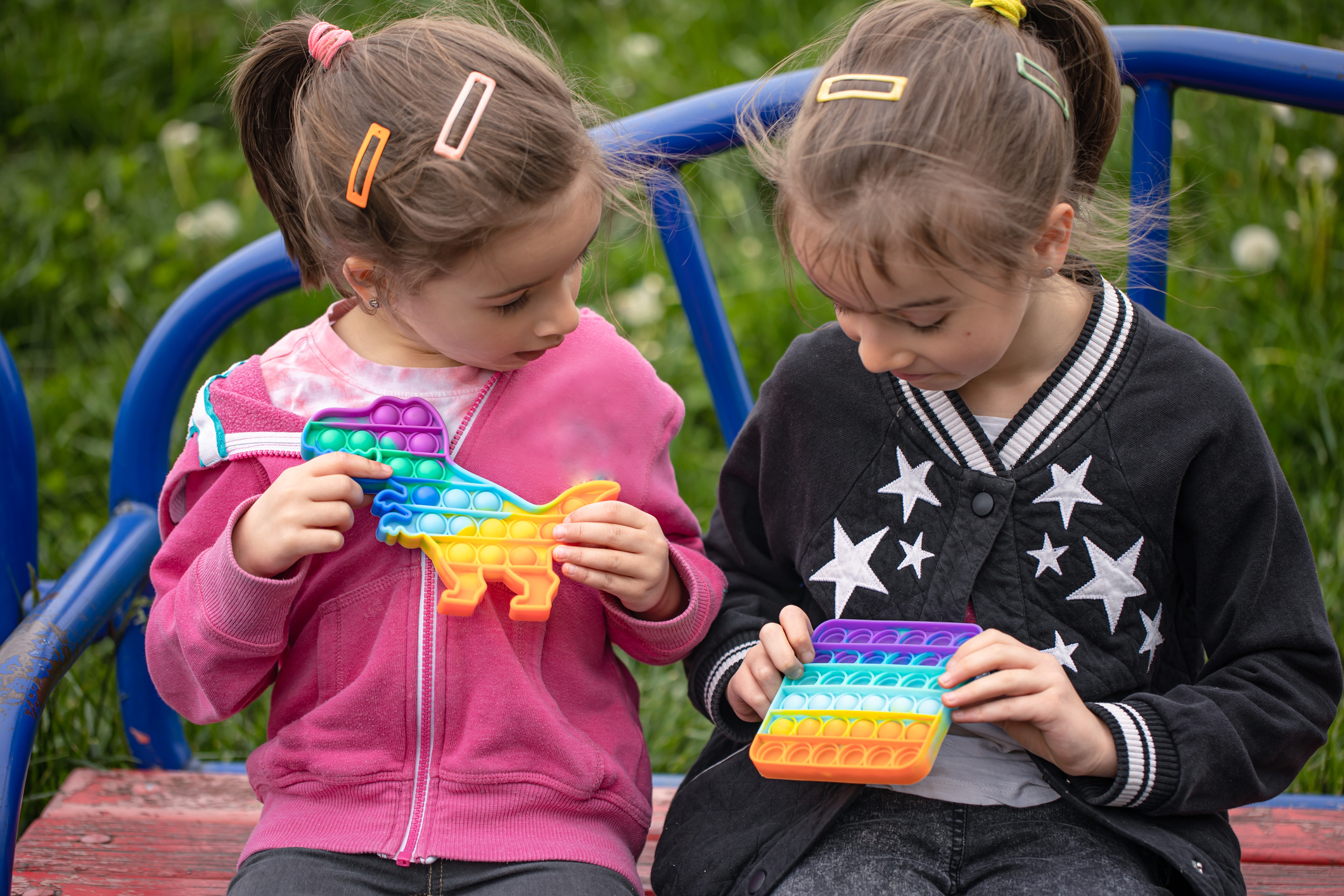 Two little girls with pale skin and colorful barettes in their light brown hair sit on a park bench playing with rainbow-colored popping fidget toys