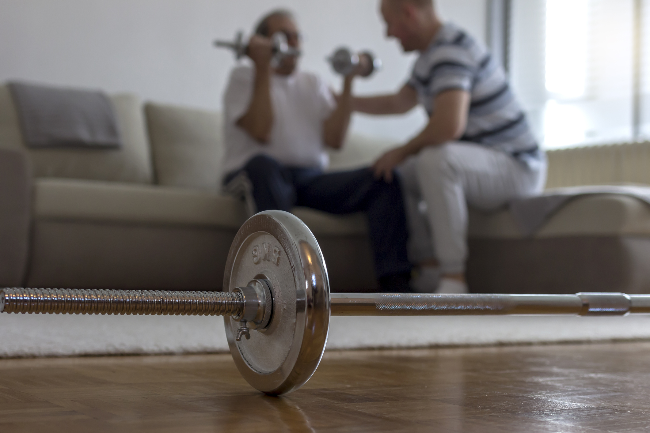 A physical therapist in white sweatpants helps an older man in loose workout clothes through arm strengthening exercises as he recovers from a stroke at home