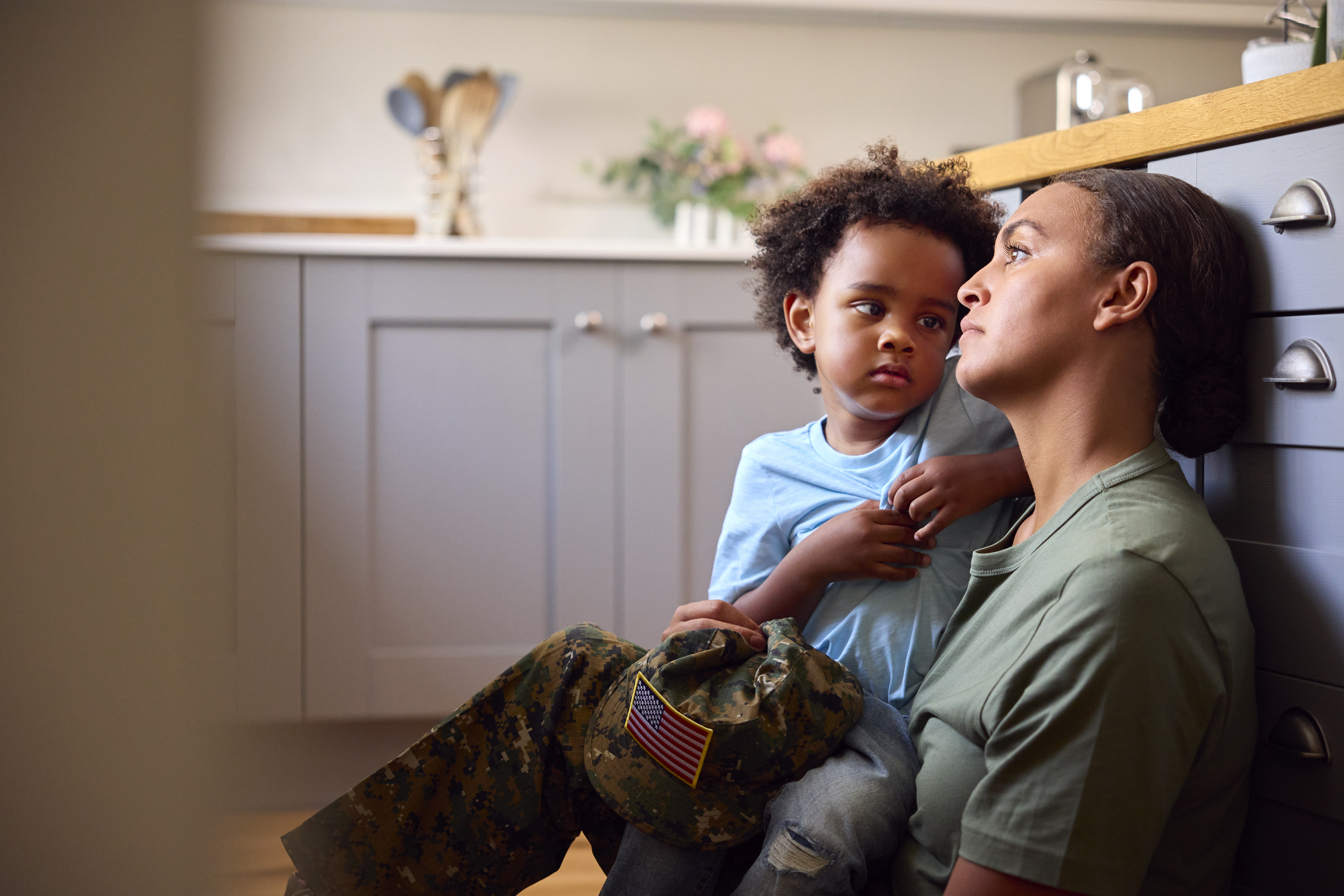 A young light-skinned Black woman wearing a military uniform with her brown hair in a tight bun sits on the floor in her kitchen, staring into space while her son, a toddler with curly black hair and a concerned expression, tries to get her attention